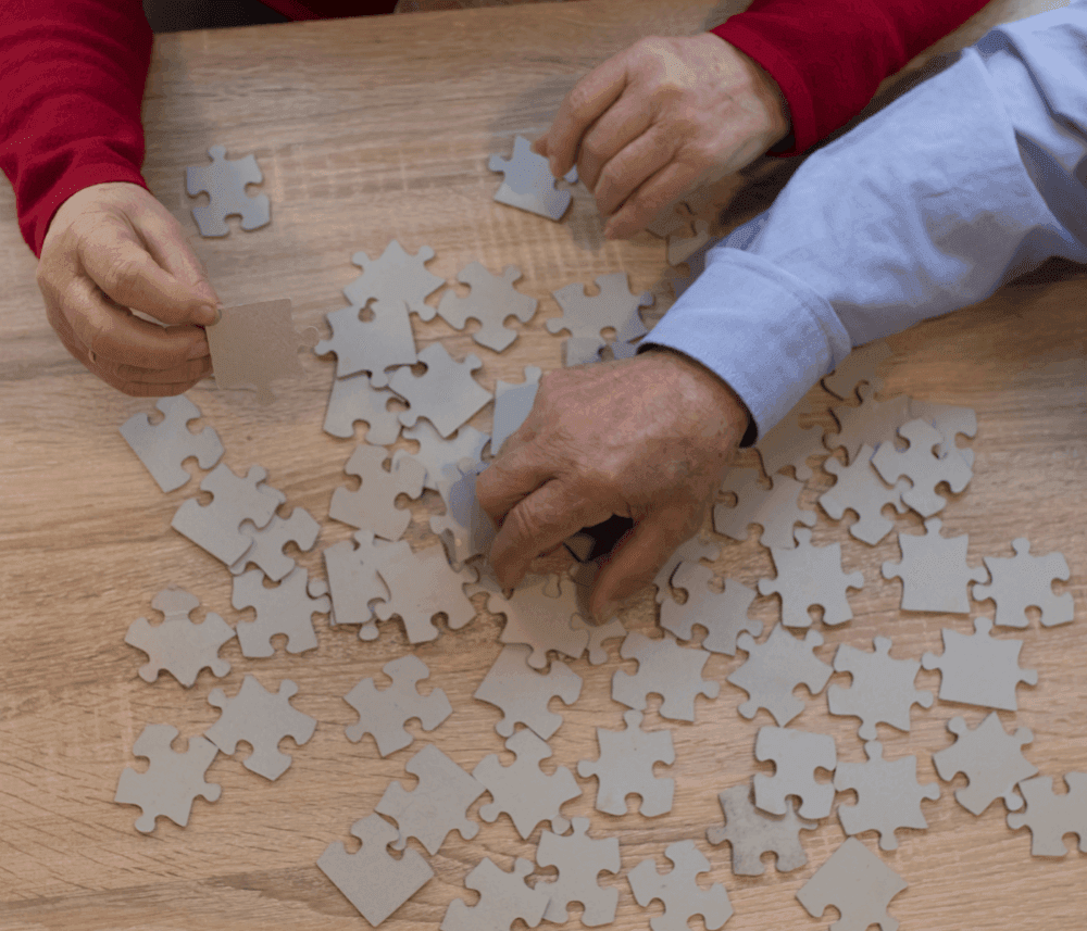 Two people sorting pieces of a blank jigsaw puzzle on a wooden table, one in a red sleeve and the other in a blue sleeve. - Home Instead