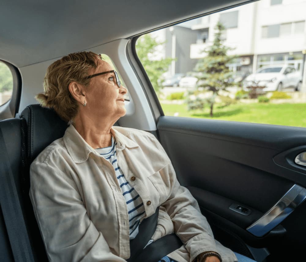 Elderly woman with glasses looking out the car window, wearing a beige jacket and striped shirt. - Home Instead