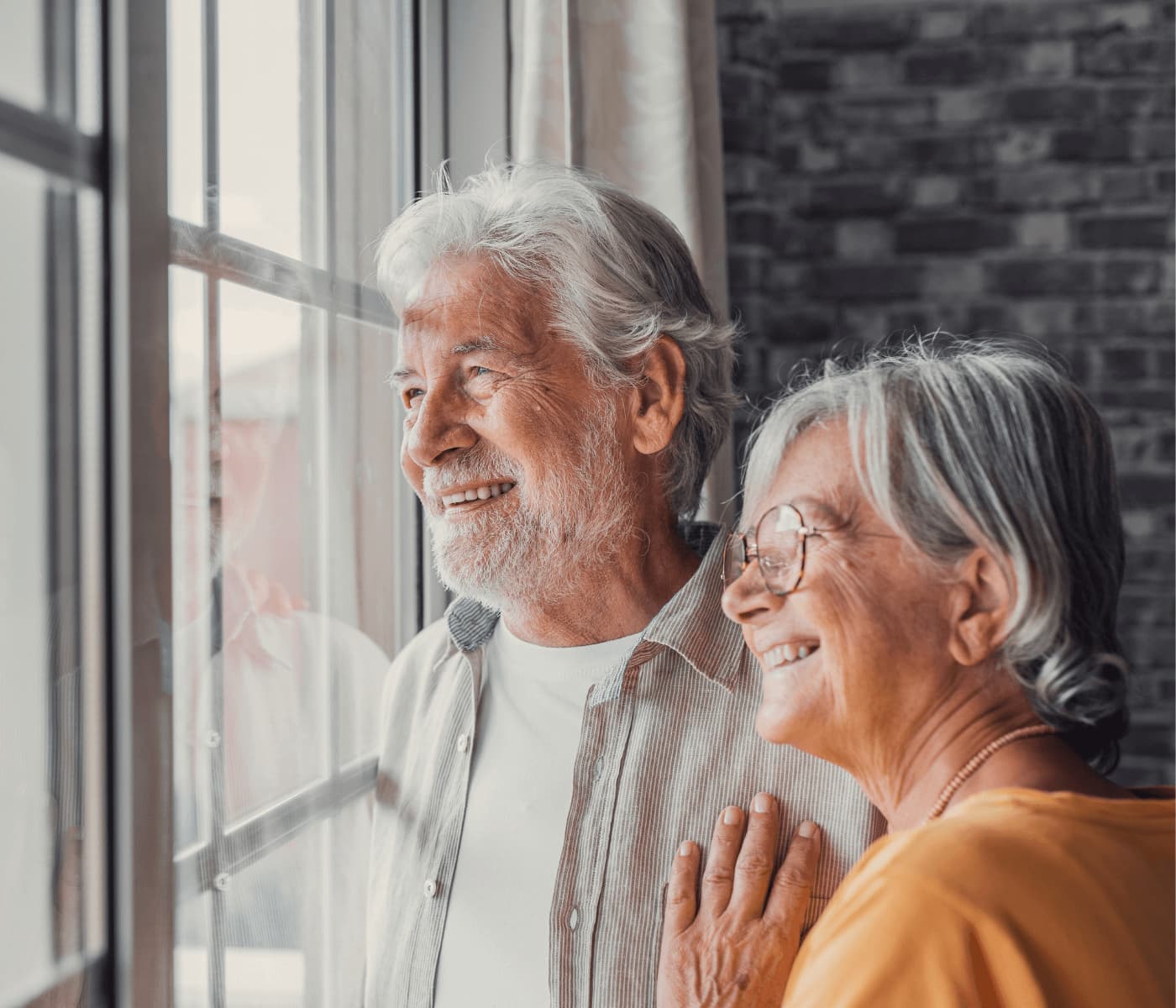 Elderly couple smiling and looking out of a window, sharing a moment of happiness indoors. - Home Instead
