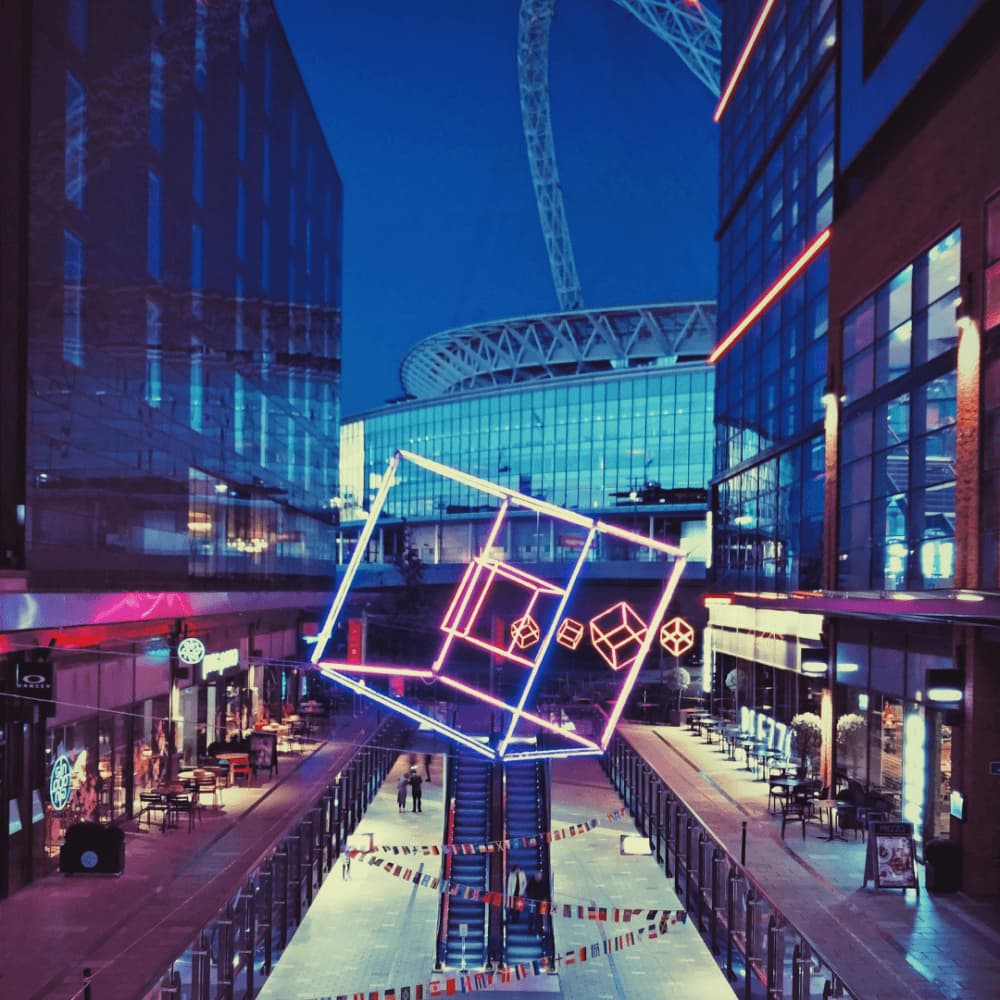 Neon-lit cube sculpture in a vibrant outdoor shopping area at night, with large stadium in the background. - Home Instead