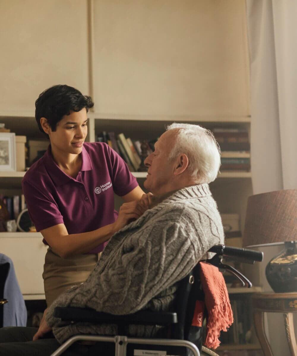 A caregiver in a purple shirt assists an elderly man in a wheelchair, tucking in his sweater inside a cozy room. - Home Instead