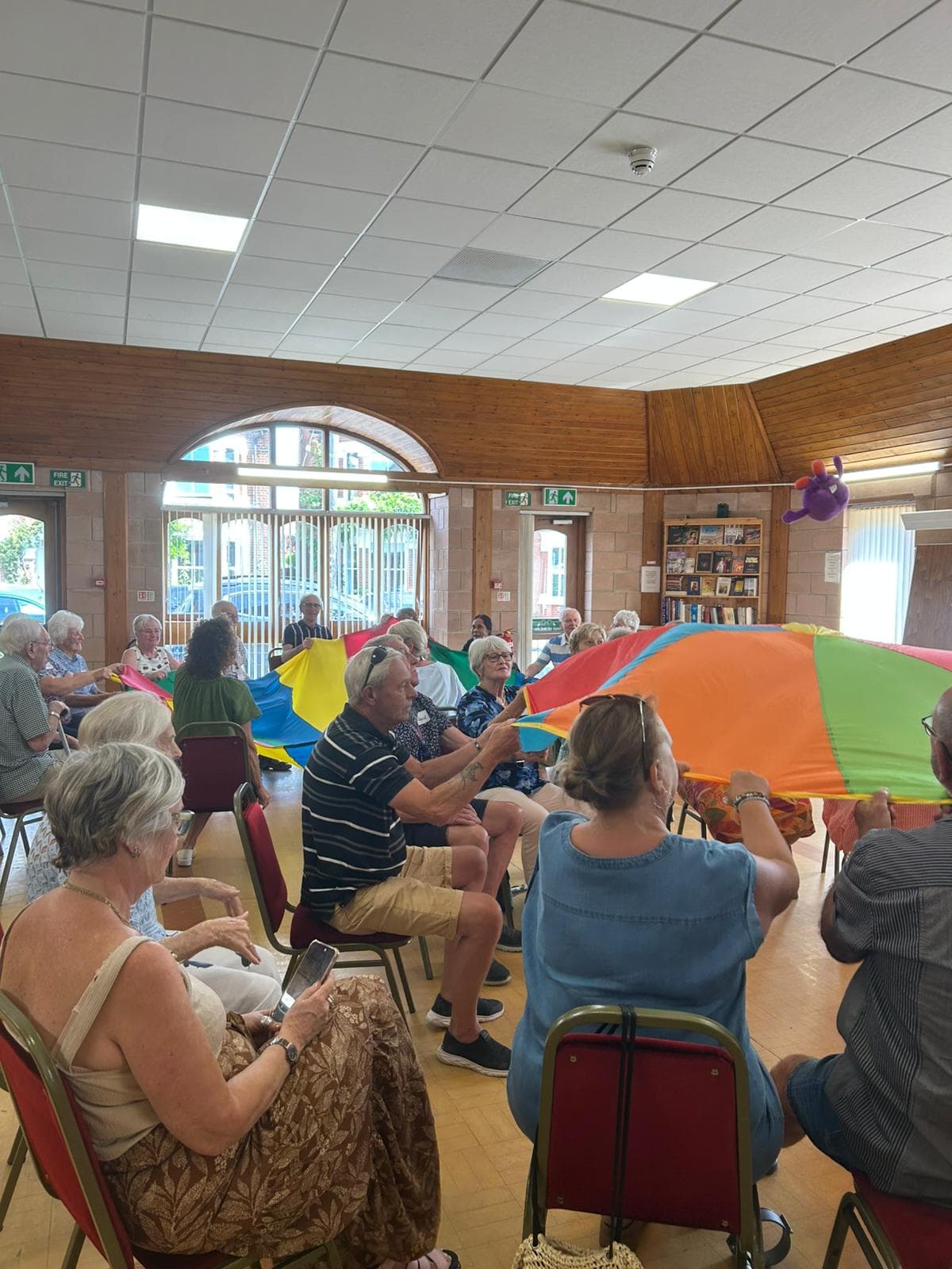 A group of older adults sit in a circle indoors, holding a colorful parachute during an activity. - Home Instead