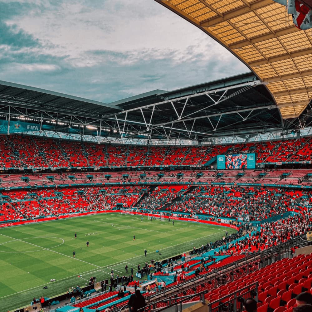 A large, open-roof stadium with a green soccer field, filled with spectators in red seats, under a cloudy sky. - Home Instead