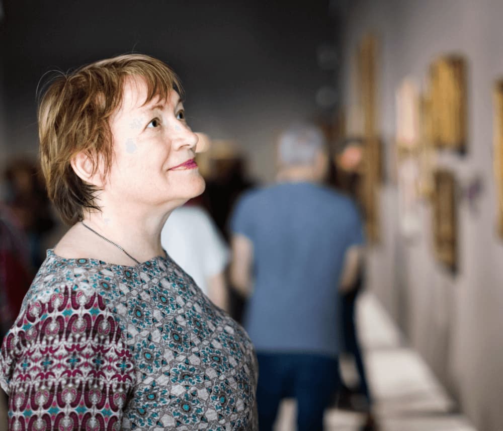 Woman in a patterned shirt looking at art in a gallery with other visitors in the background. - Home Instead