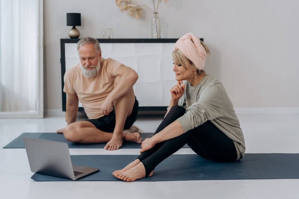 Two people sitting on yoga mats indoors, looking at a laptop screen. Background includes light-colored walls and decor. - Home Instead