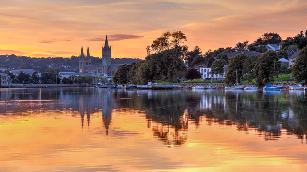 Sunset over a calm river with boats docked and Truro cathedral with twin spires in the background, trees reflected in the water. - Home Instead