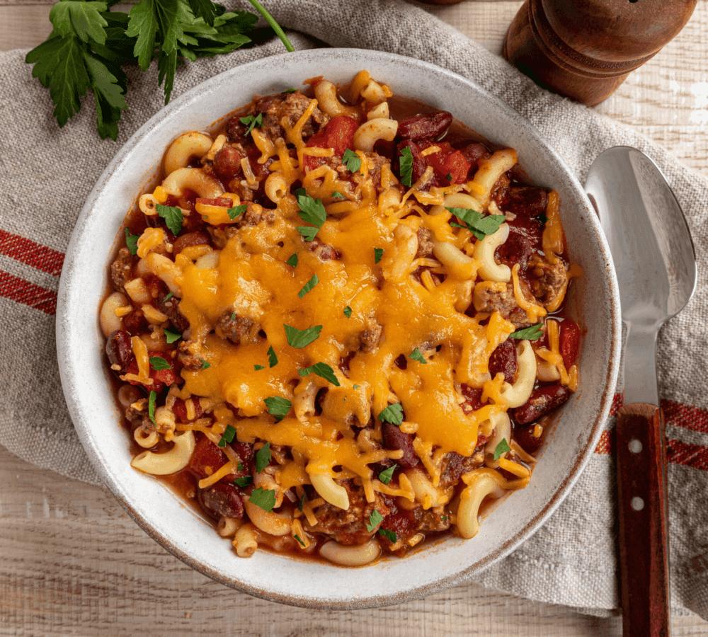 A bowl of chili mac with melted cheese on top, garnished with parsley, next to a spoon and fresh parsley on a cloth. - Home Instead