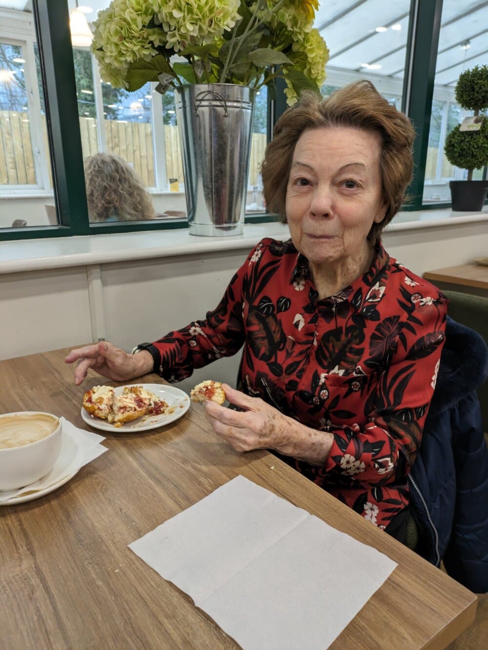 Elderly woman in a floral shirt enjoys pastries at a cafe table, surrounded by plants and windows in the background. - Home Instead