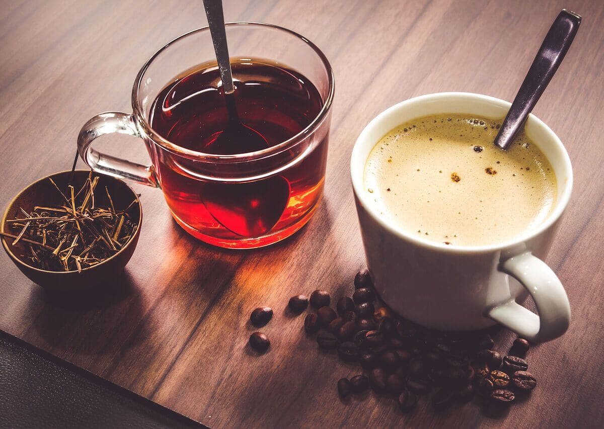 Cup of tea next to loose tea leaves in a small bowl, and a cup of coffee with coffee beans scattered around it, on a wooden table. - Home Instead
