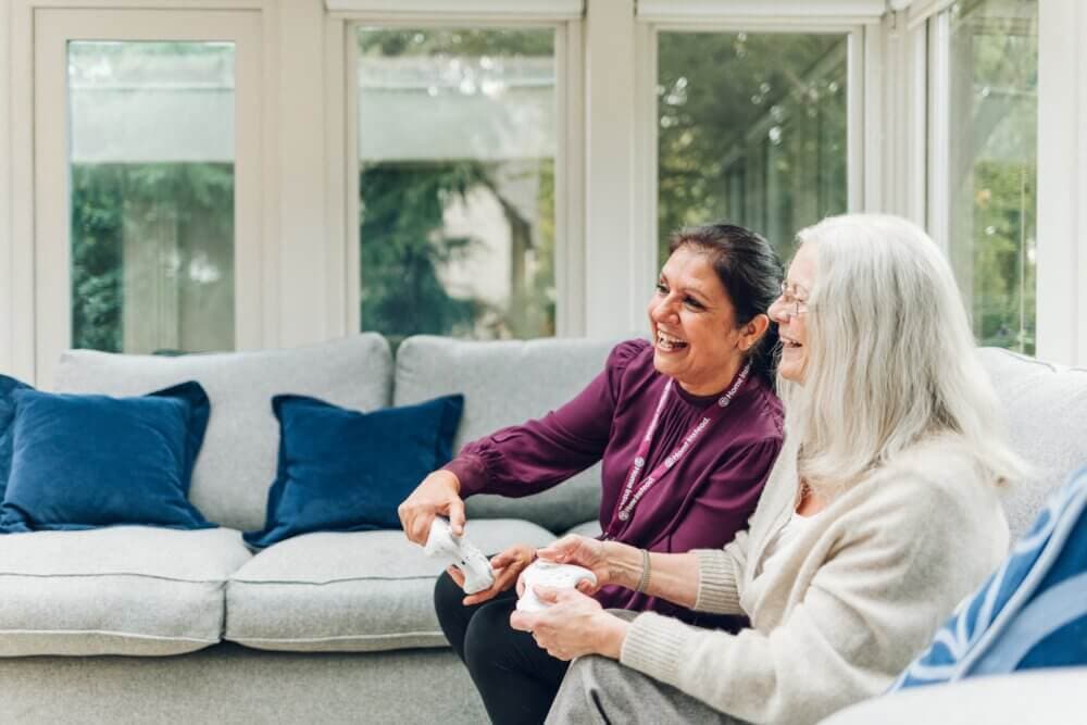 Two women, one elderly and one middle-aged, enjoying playing video games on a couch in a bright living room. - Home Instead