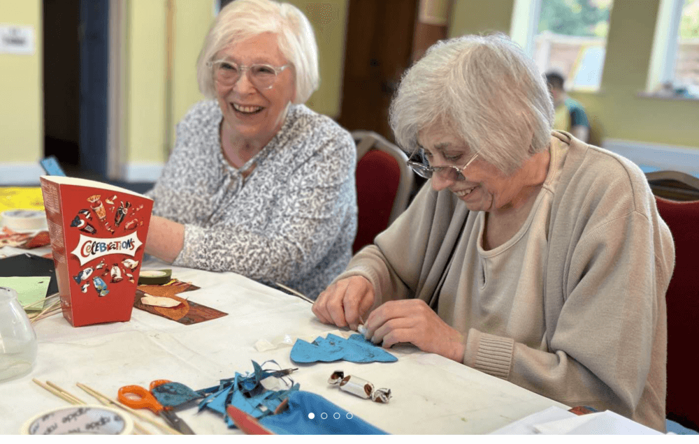 Two elderly women sitting at a table, crafting and smiling, with a box of chocolates nearby. - Home Instead