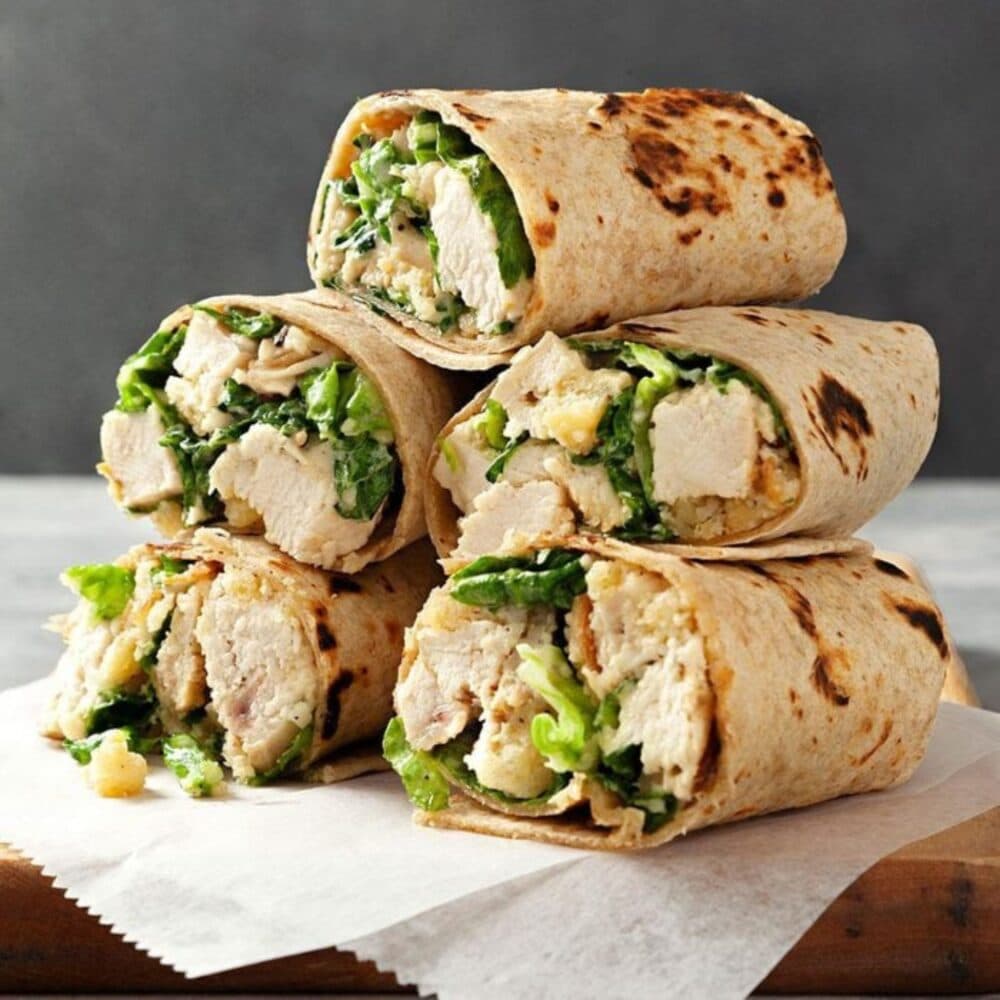 Four chicken Caesar wraps stacked on a piece of parchment paper, filled with chicken, lettuce, and croutons. - Home Instead Renfrewshire and Barrhead home care services - meal preparation.