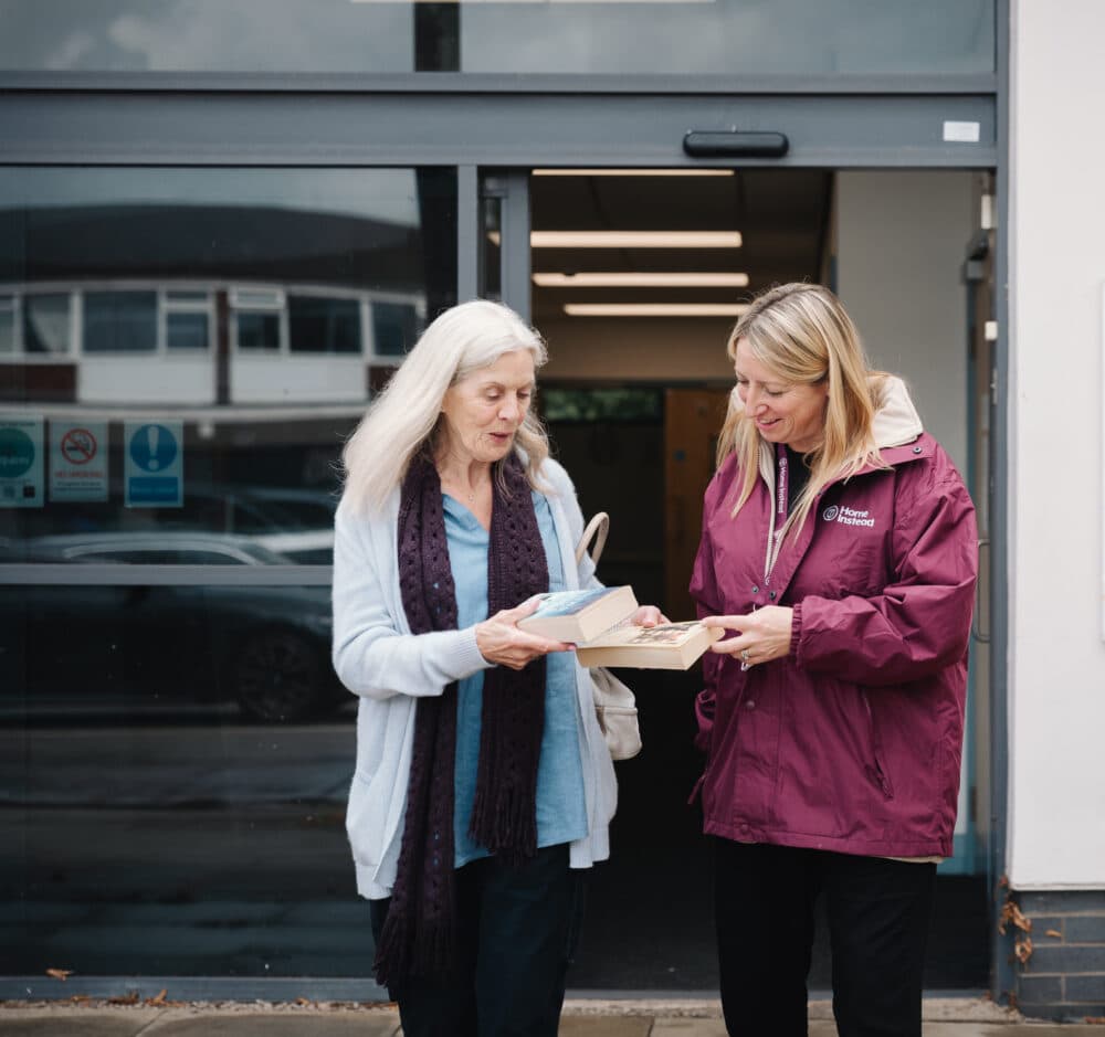Two women standing outside a building, one handing a box to the other, both smiling and engaged in conversation. - Home Instead