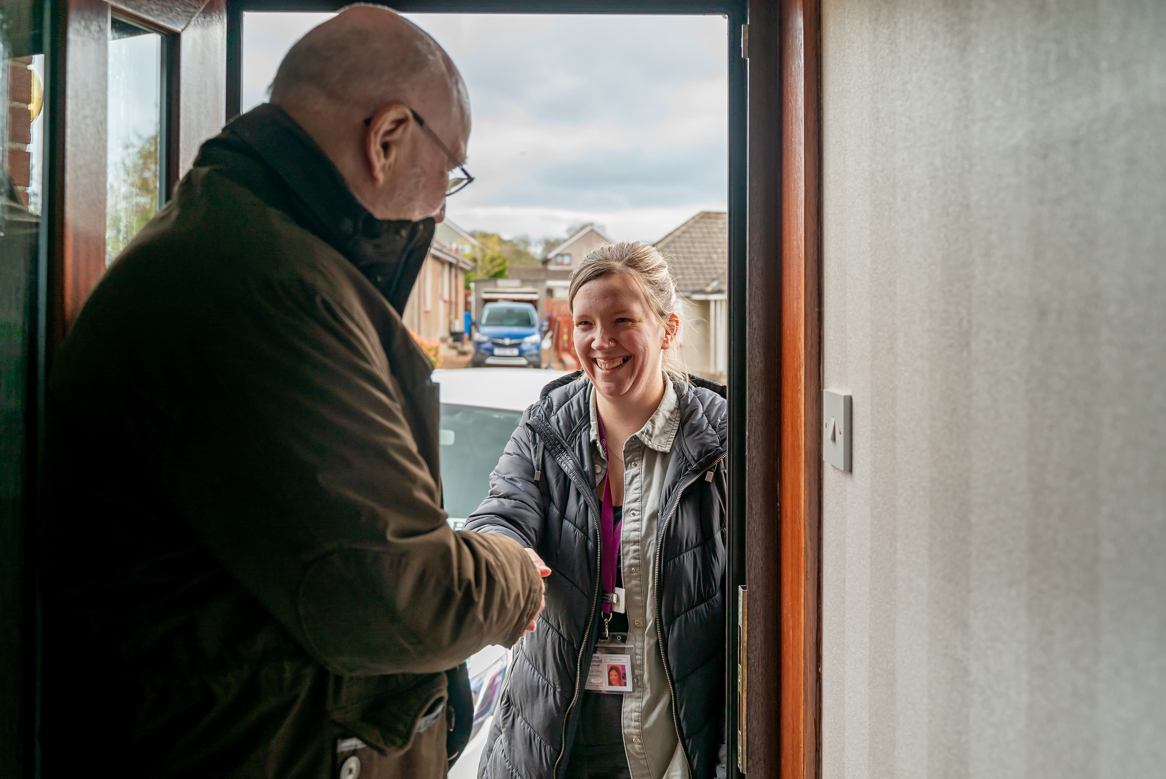 Person at door shaking hands with a smiling visitor wearing a coat and lanyard, standing on a residential street. - Home Instead