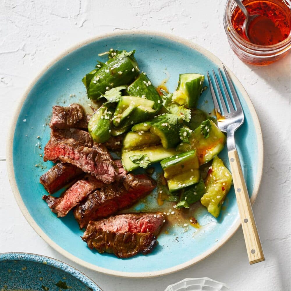 A plate with sliced steak, cucumber salad, a fork, and a jar of sauce. - Home Instead Renfrewshire and Barrhead home care services - Meal Preparation.