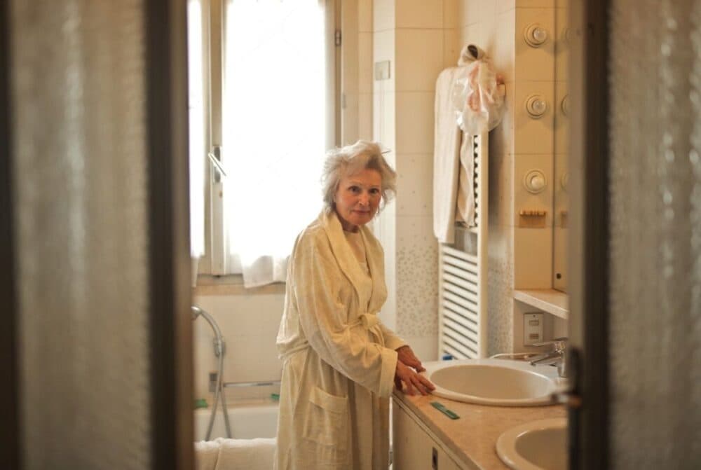 An elderly woman wearing a robe stands in a bathroom, leaning on the sink and looking toward the camera. - Home Instead