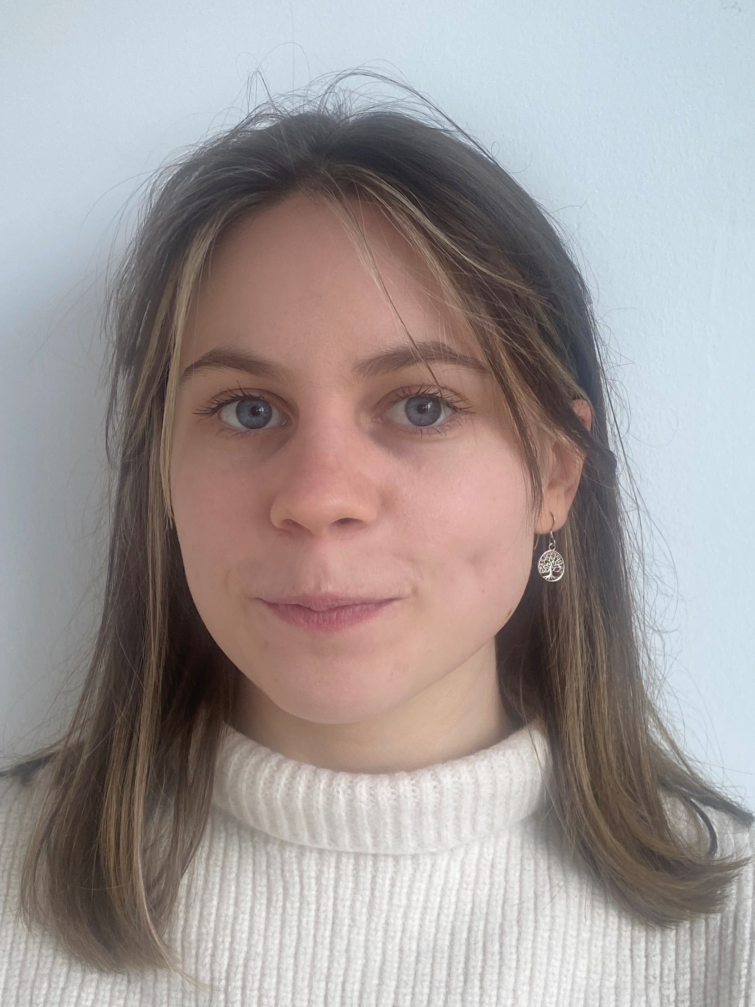 Young woman with long brown hair and earrings smiles slightly against a light background, wearing a white turtleneck. - Home Instead