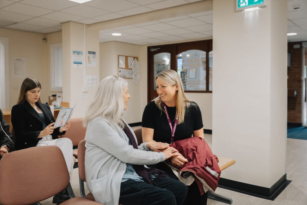 A woman comforts an older woman seated in a waiting area while another person reads a pamphlet in the background. - Home Instead