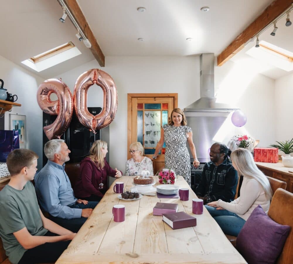 a family sitting on chairs with long table and with a 90 number balloon with mugs and cake on top of the table, everyone is smiling and happy inside the kitchen
