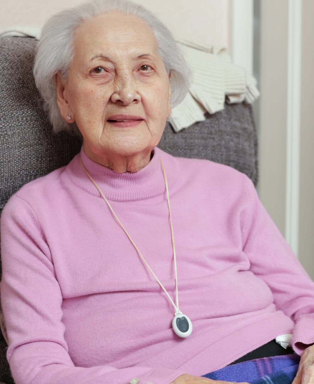 Elderly woman in a pink sweater sitting on a chair, wearing a medical alert pendant around her neck. - Home Instead