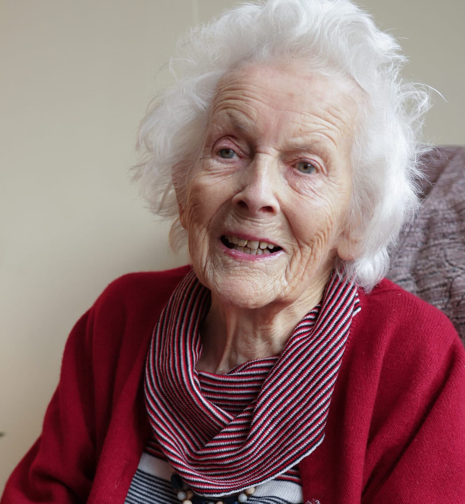 Smiling elderly woman with white hair wearing a red sweater and striped scarf, seated indoors. - Home Instead