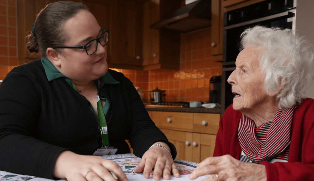 A younger woman chats with an elderly woman at a kitchen table, both appearing engaged in conversation. - Home Instead