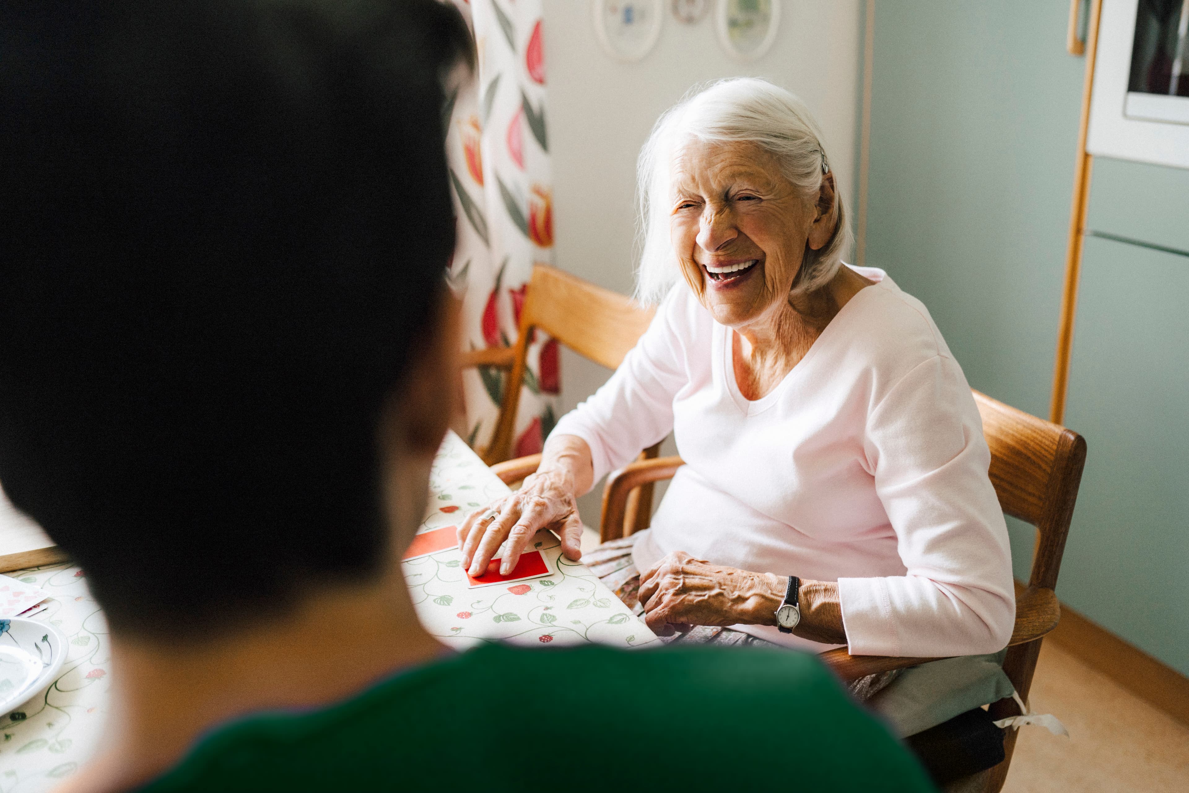 Smiling elderly woman sits at a table, talking to someone whose back is to the camera. - Home Instead