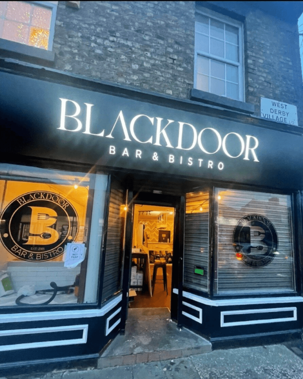 Blackdoor Bar & Bistro exterior with illuminated sign and open door, seen at dusk on a city street. - Home Instead
