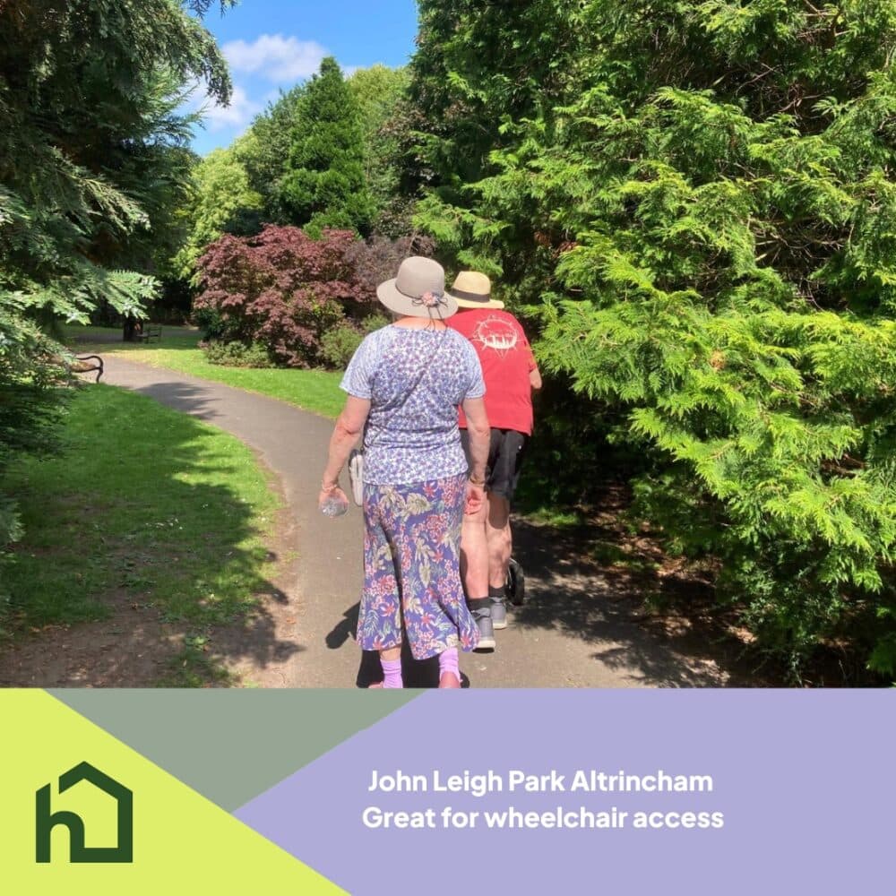 Two people walk on a paved path through a green park, with trees and bushes on both sides. - Home Instead