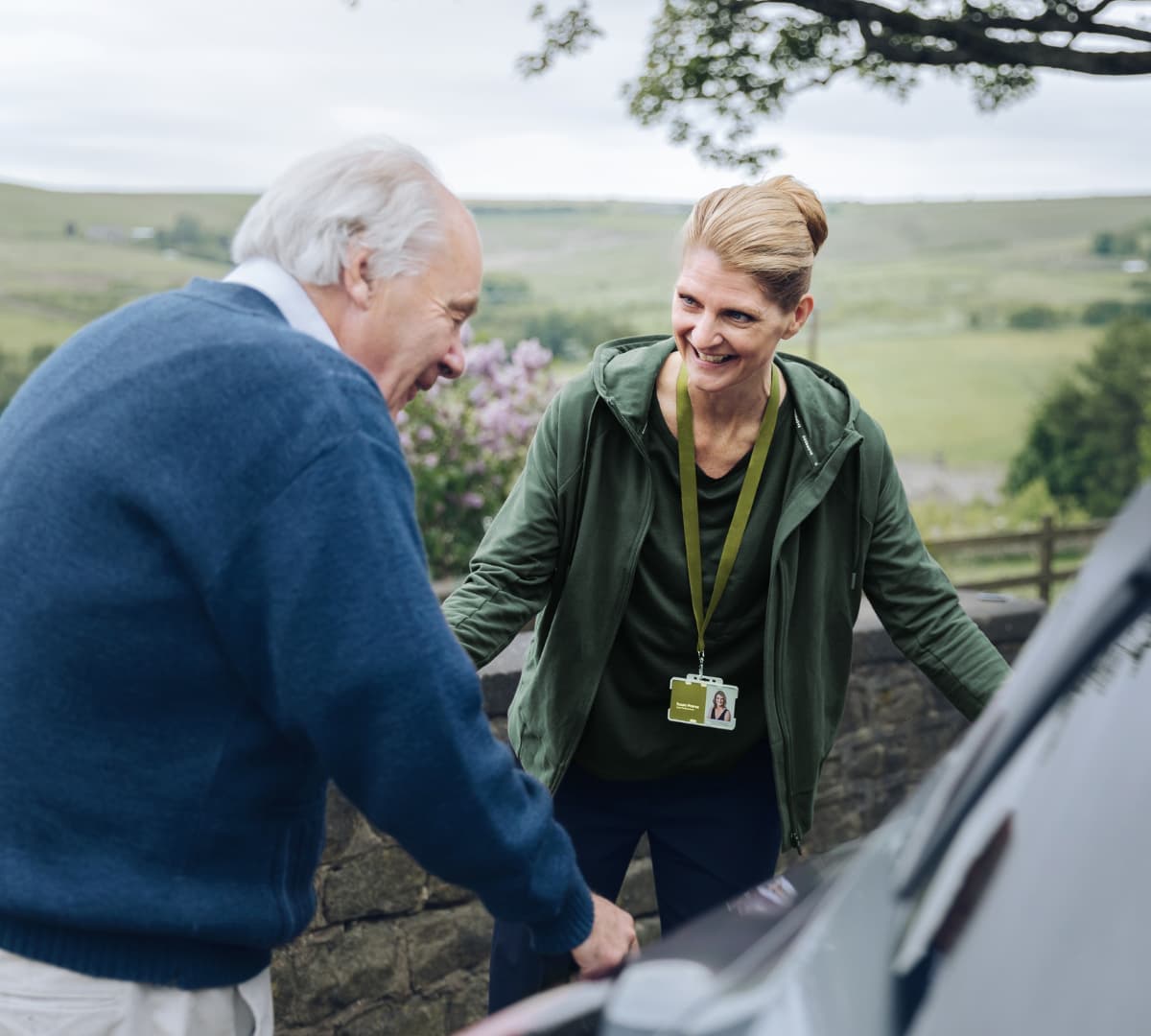 An older male adult wearing blue pullover sweater and with grey hair riding a car with the help of his young female carer wearing green sweater smiling while helping