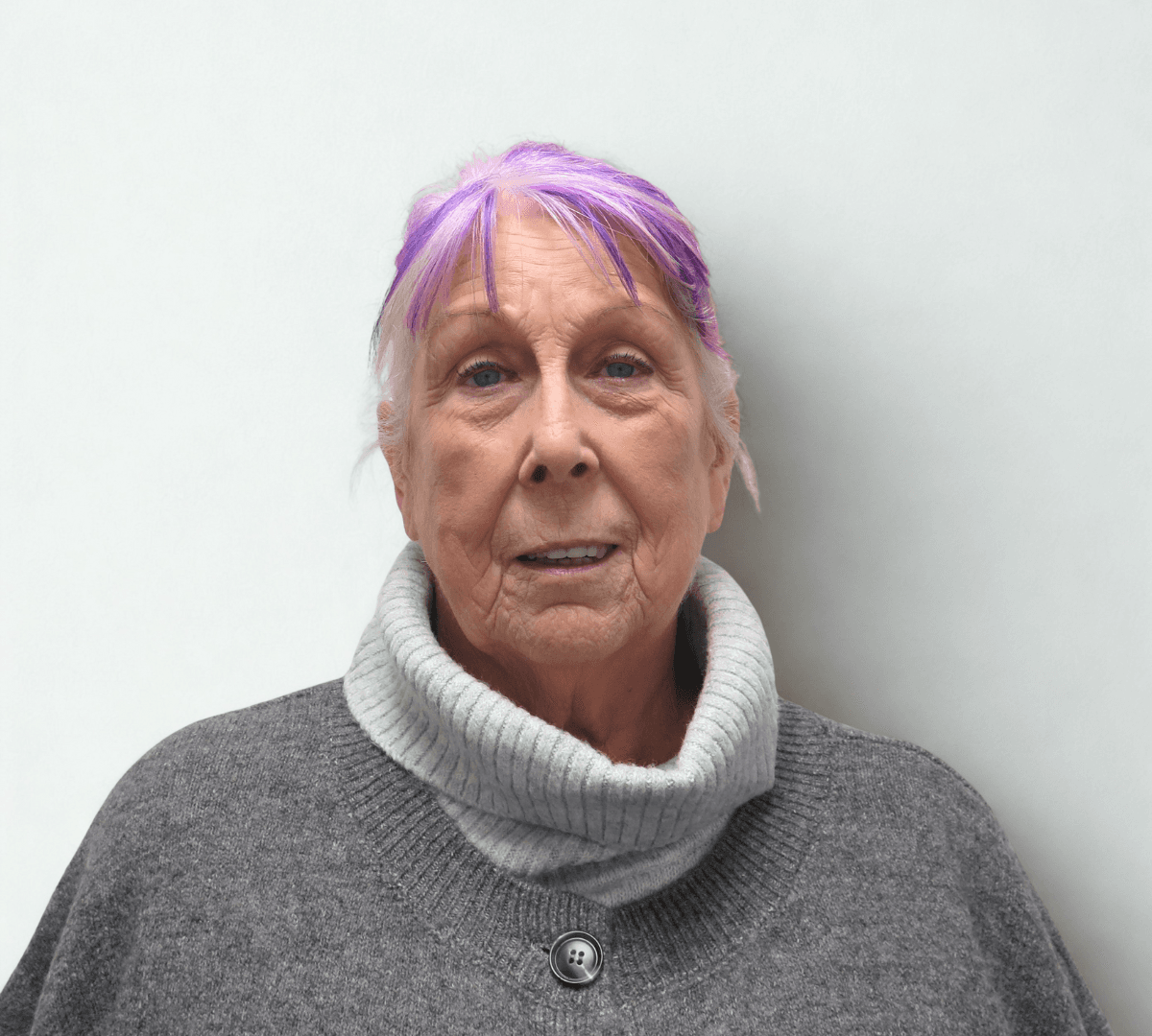 Older woman with light purple hair and a grey turtleneck sweater, standing against a plain light background. - Home Instead
