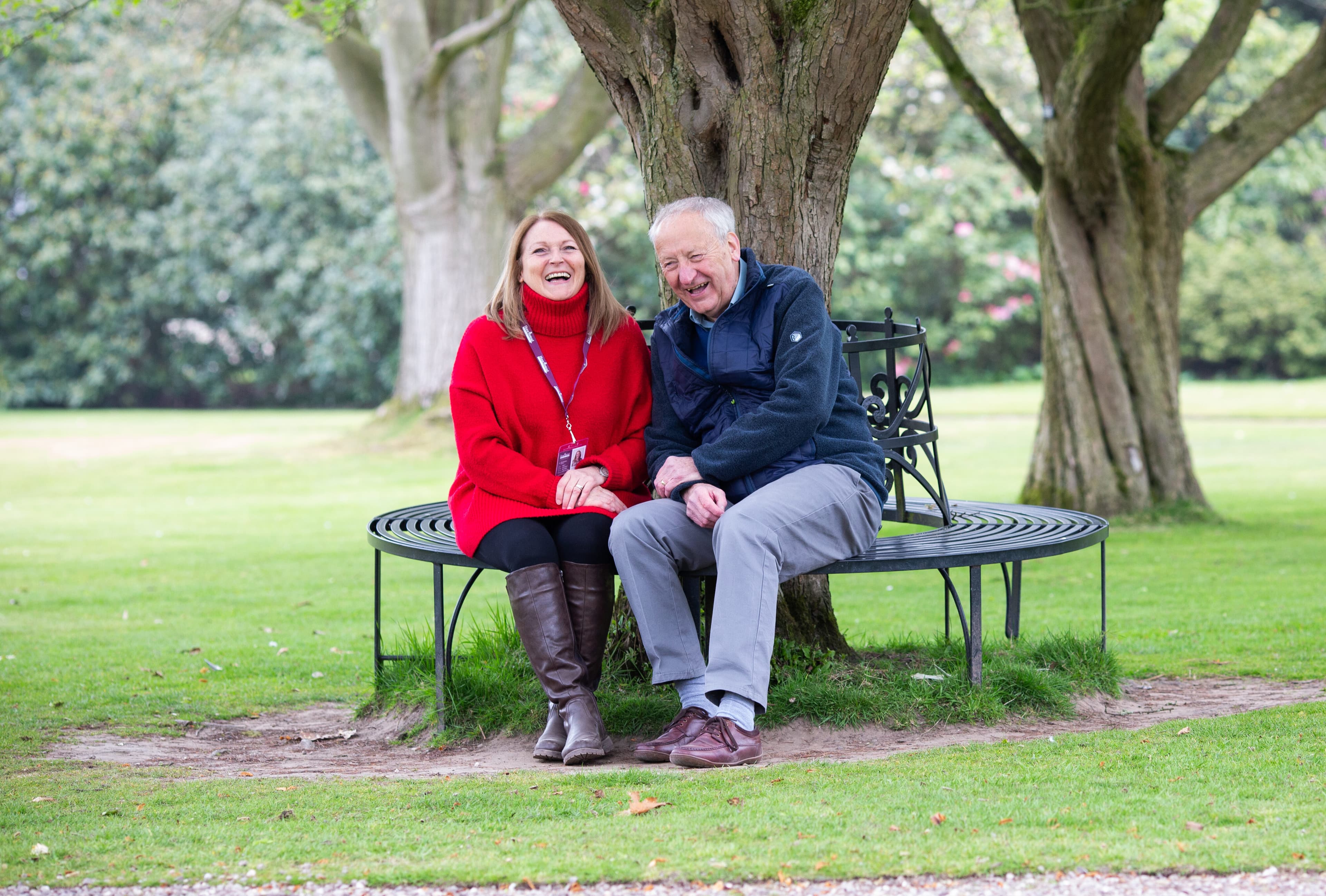 Elderly man and his female carer sitting on a circular bench around a large tree in a park, looking happy