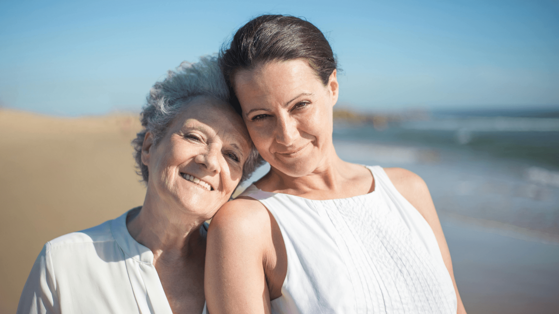 Smiling older woman and her adult daughter standing together on a sunny beach, sharing a warm moment. Ideal image for Home Instead West Exeter, Teignbridge and South Devon, representing compassionate respite care and family support.