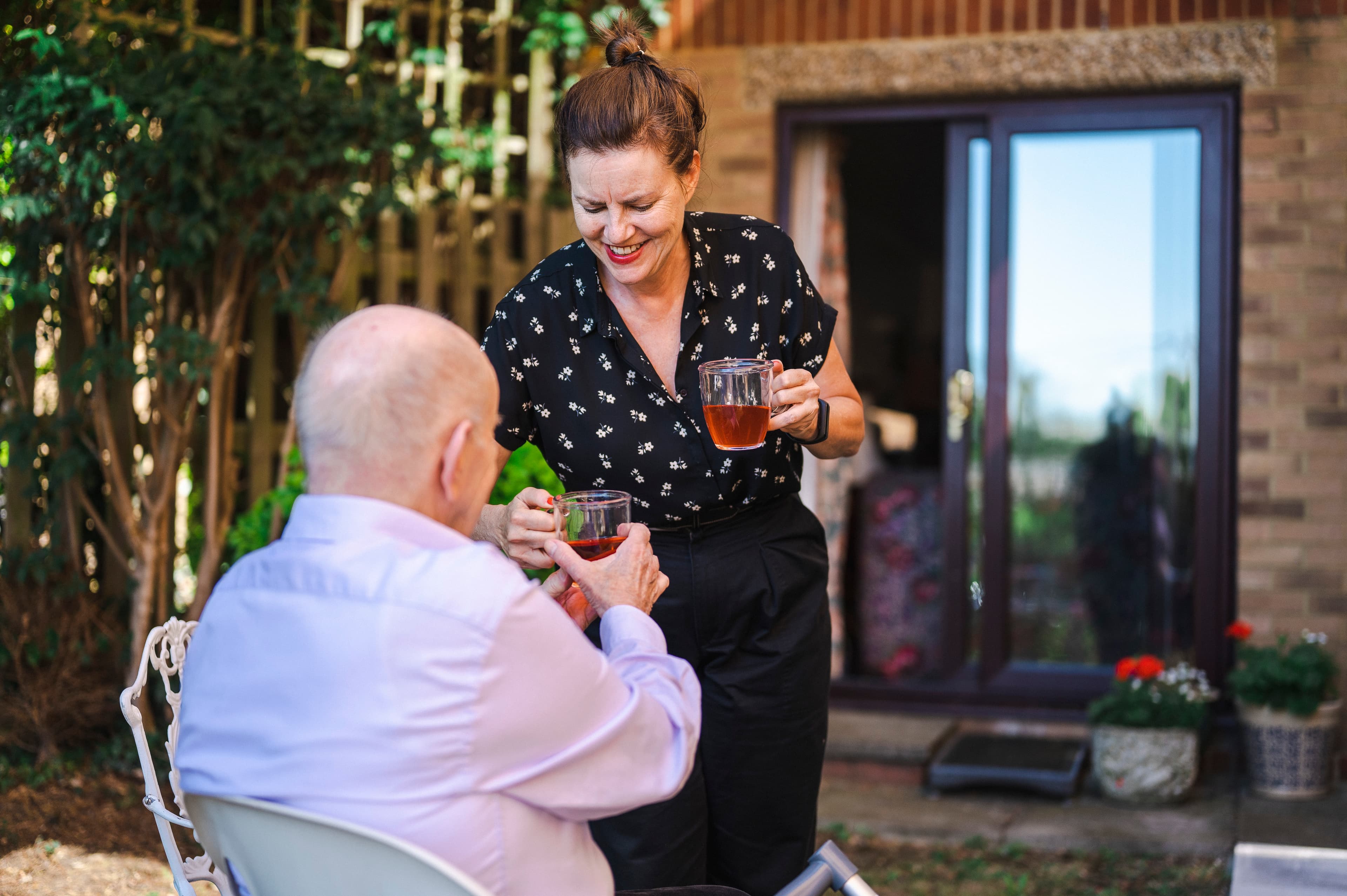 Smiling woman hands a mug of tea to an older man sitting outside in a garden near a house. - Home Instead