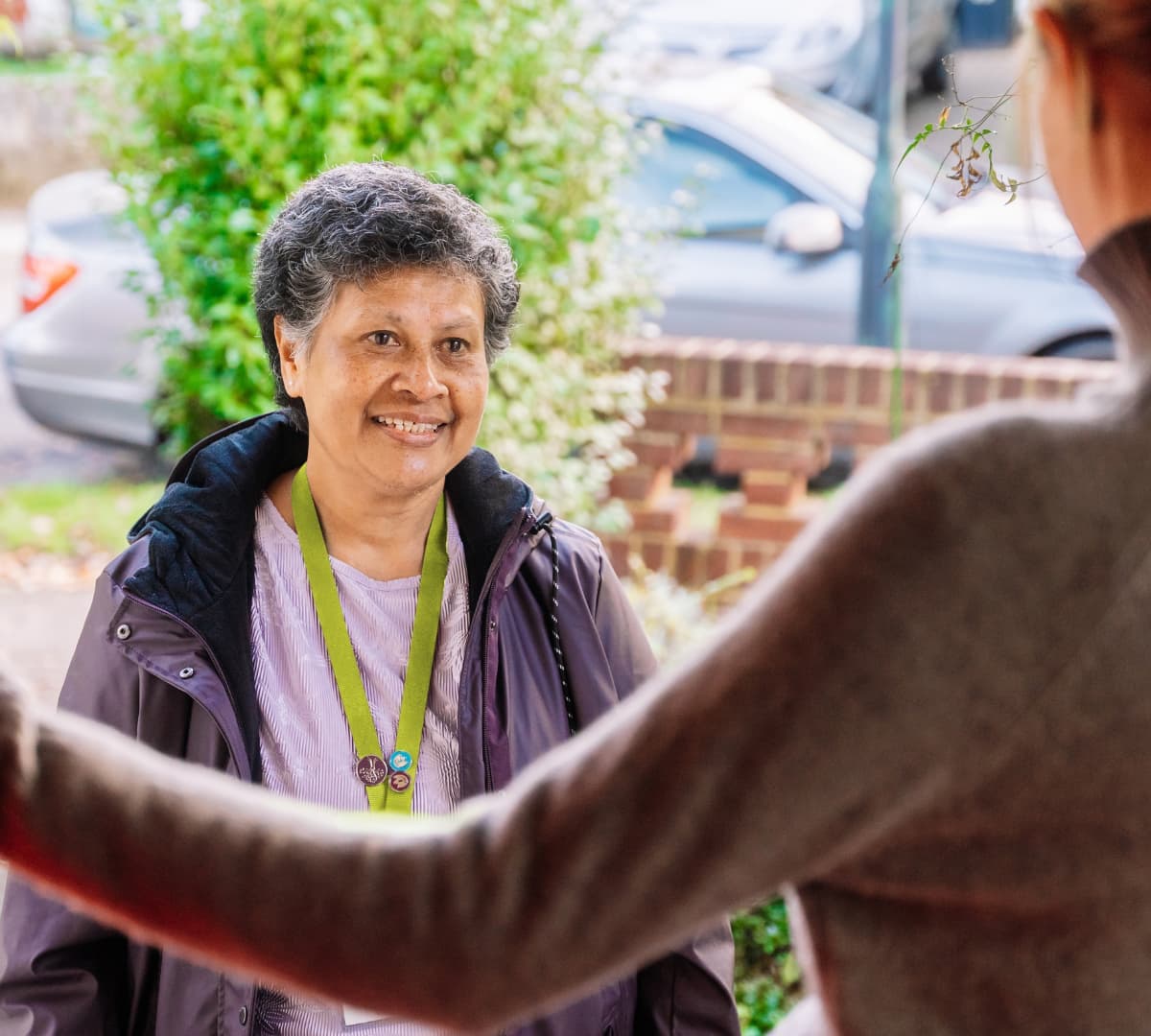 Smiling woman wearing a green lanyard stands outside, greeting another person at a doorway. - Home Instead