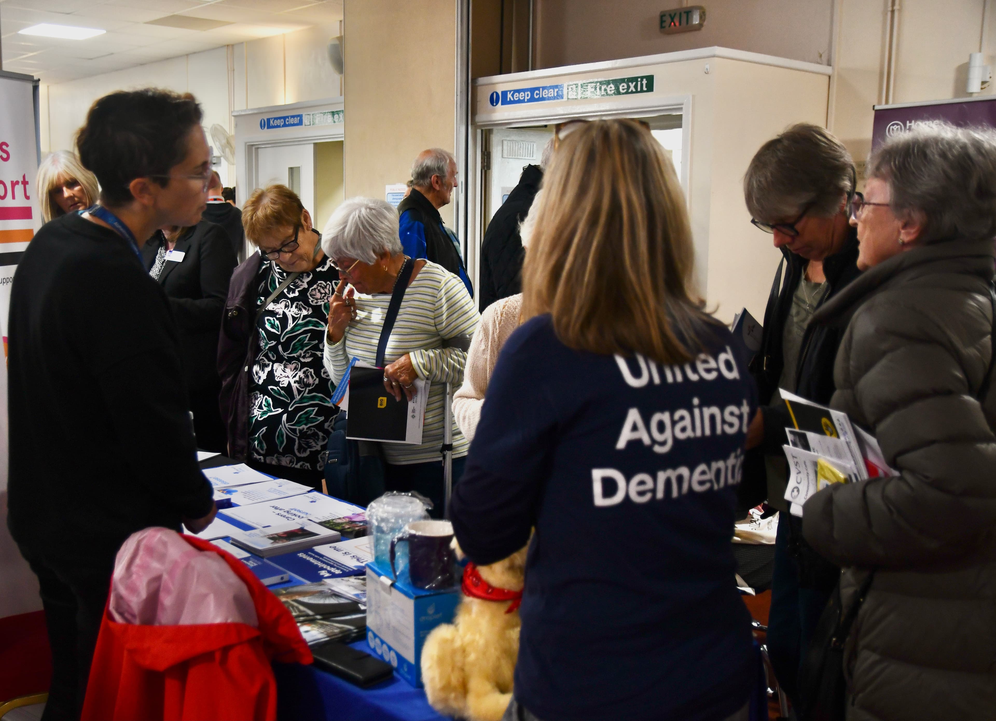 People gather around an information table at an indoor event; one person wears a "United Against Dementia" shirt. - Home Instead