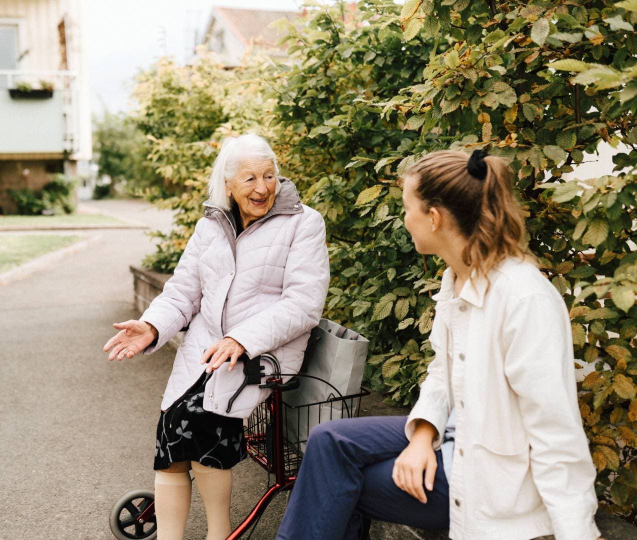 An elderly woman with a walking frame smiles and talks with a younger woman on a pavement near greenery. - Home Instead