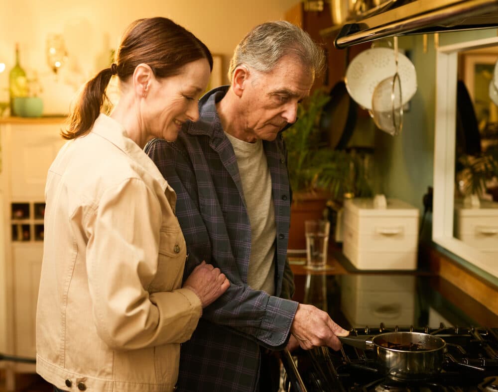 An older couple stand together while cooking at a cooker in a warmly lit kitchen. - Home Instead
