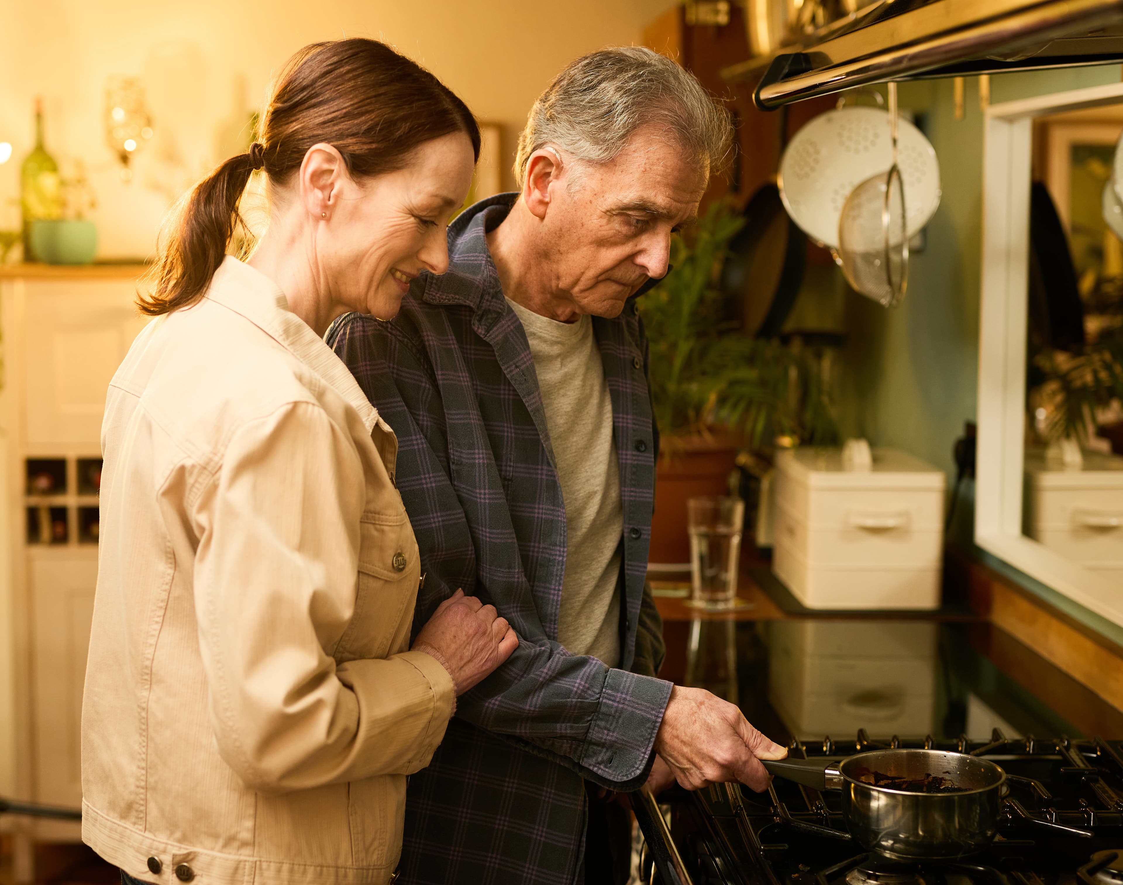 An older couple stand together while cooking at a cooker in a warmly lit kitchen. - Home Instead