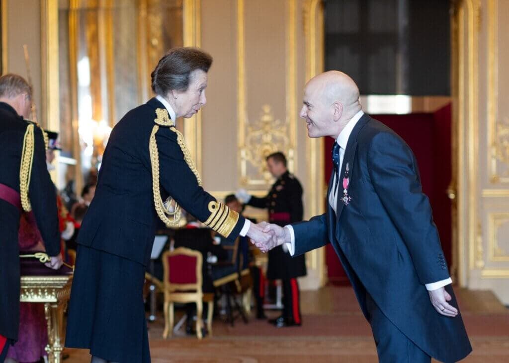 A woman in formal military uniform shakes hands with a man in a suit at an ornate ceremony. - Home Instead