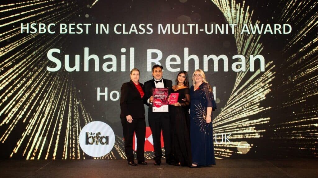 Four people stand on stage holding awards in front of a display reading "HSBC Best in Class Multi-Unit Award Suhail Rehman. - Home Instead