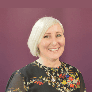 Smiling woman with short blonde hair in a floral top, posing in front of a purple background. - Home Instead
