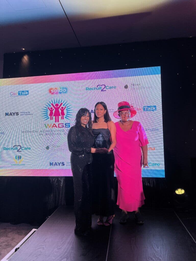 Three women from Home Instead Greenwich & Bexley celebrate double success at the WAGS Awards, standing on stage holding an award, with sponsor logos displayed on a screen behind them. - Home Instead
