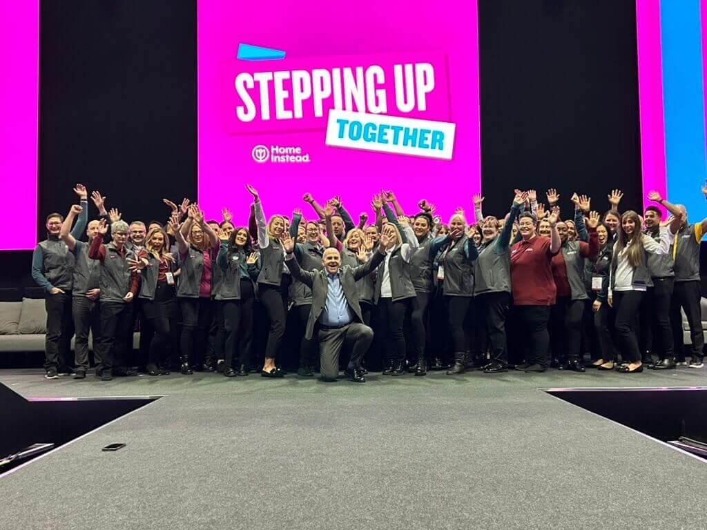 A large group of people smiling and waving on stage under a sign that reads "Stepping Up Together". - Home Instead