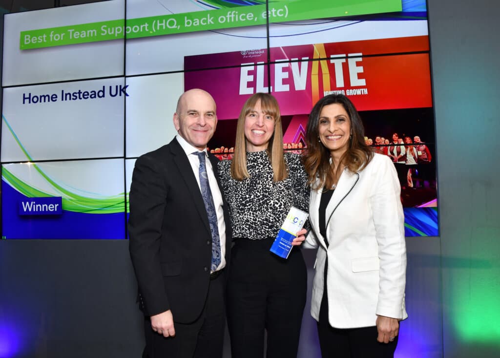 Three people smiling at an awards event, holding a trophy, with a screen behind showing "Home Instead UK Winner" — celebrating multiple award wins for Home Instead at the Home Care Awards. - Home Instead