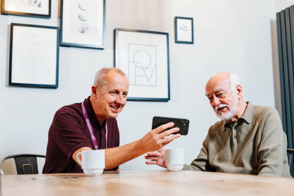 Two older men sit at a table with coffee, smiling and looking at a mobile phone together. - Home Instead