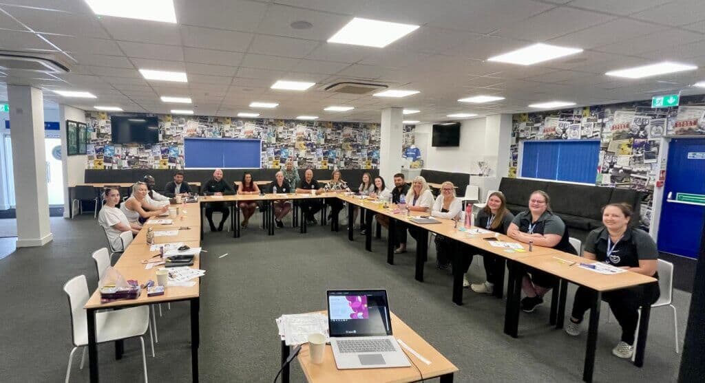 A group of people sit around a large U-shaped table in a well-lit meeting room with posters on the walls. - Home Instead
