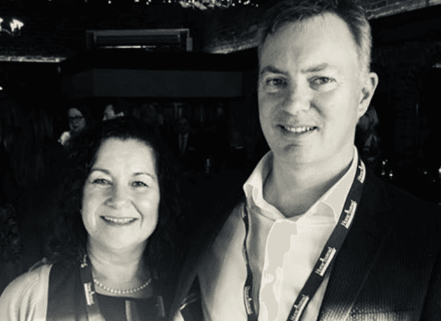 Cath & Mike Chalton, featured in the Case Study Home Instead Franchisees, smile at an event, both wearing name badges, in a dimly lit room. - Home Instead