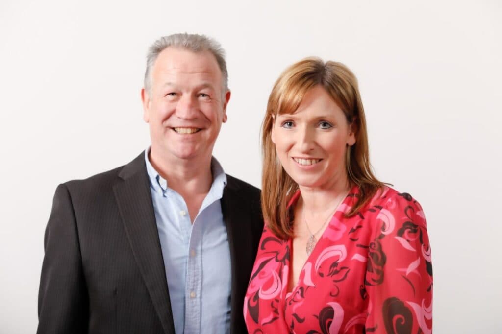 A smiling man in a suit and a woman in a pink patterned top pose together against a white background. - Home Instead