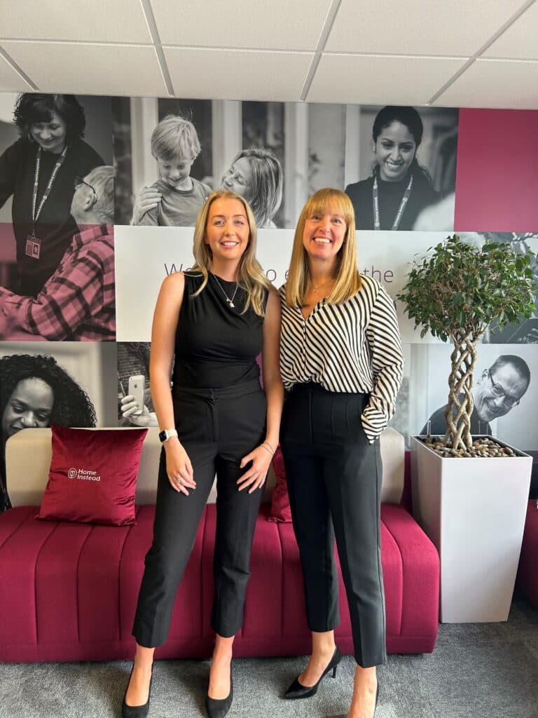 Two women smiling, standing in front of a maroon settee and a wall with black-and-white photo portraits. - Home Instead